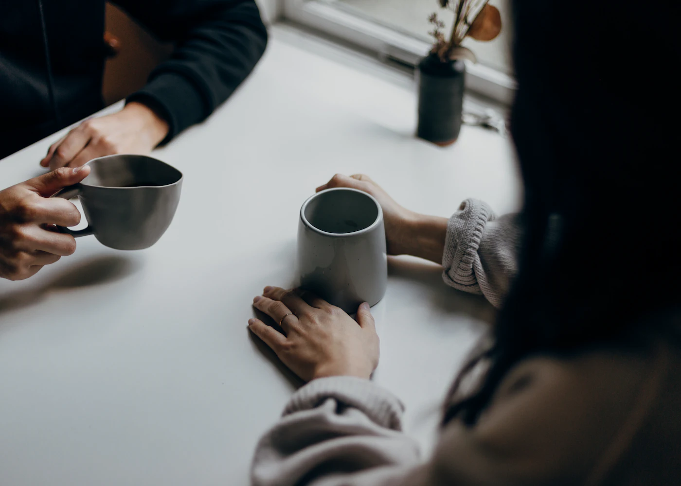 Two people talking across a white table, each holding a grey mug